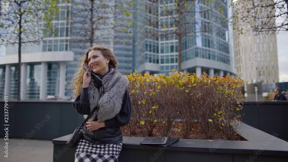 joyful young woman is talking by phone standing outdoors in modern area of city in fall day joyful young woman is talking by phone standing outdoors in modern area of city in fall day