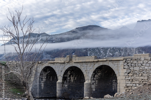Old bridge in ruins that is generally flooded by the waters of the reservoir of Riaño in Leon, Spain. In the background you see the mountains between the morning mists. There is a tree near the bridge photo
