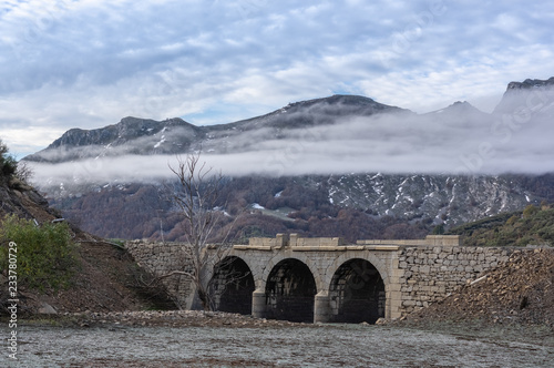 Old bridge in ruins that is generally flooded by the waters of the reservoir of Riaño in Leon, Spain. In the background you see the mountains between the morning mists. There is a tree near the bridge photo