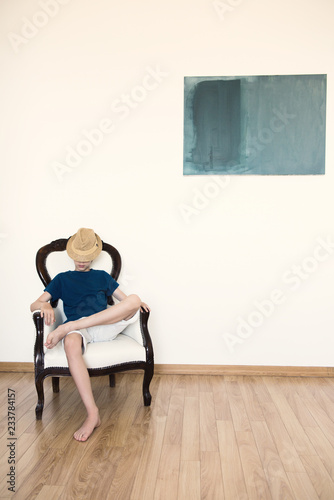 Boy sitting in armchair in empty room with white walls. Abstract accommodation. Minimalism in lodging, image toned.