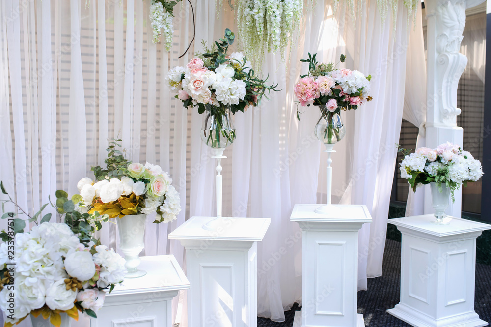 Fototapeta premium Closeup photo of white pillars with vases with flowers on them and a part of an wedding arch with white curtains on background