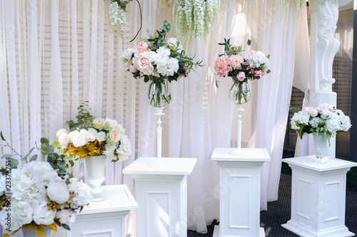 Closeup photo of white pillars with vases with flowers on them and a part of an wedding arch with white curtains on background © Med Photo Studio