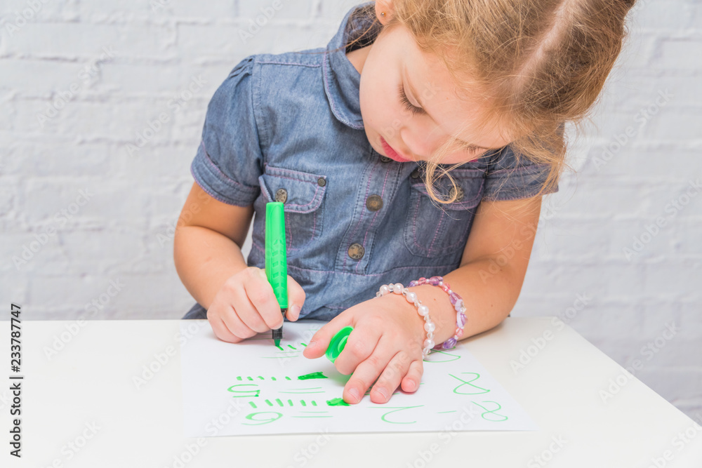 a child, a girl at the table writes, draws on a piece of paper, against a white brick wall a child, a girl at the table writes, draws on a piece of paper, against a white brick wall