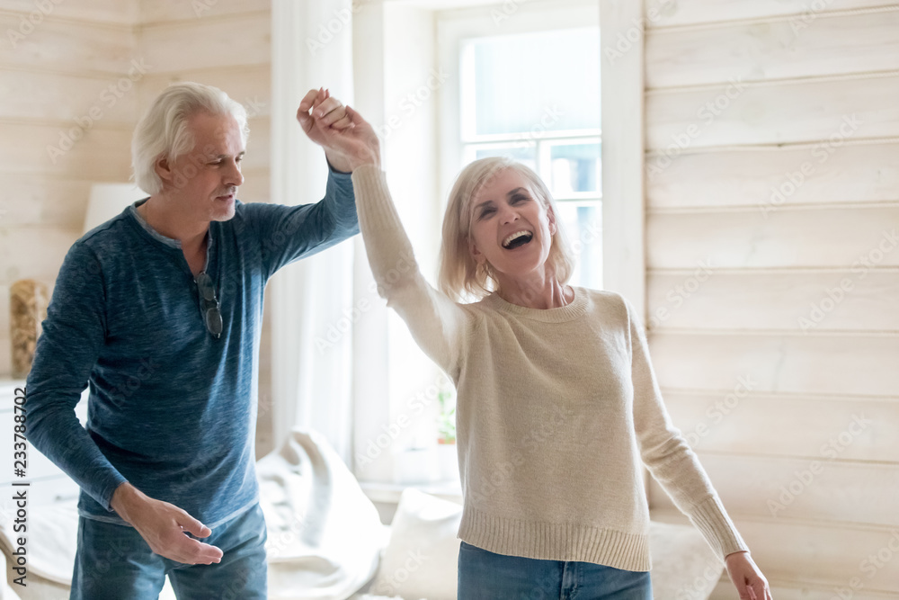 Fototapeta premium Happy senior husband and wife have fun spending time in country house together, smiling aged couple dance swirling and swaying at home, excited elderly man and woman waltz turning around