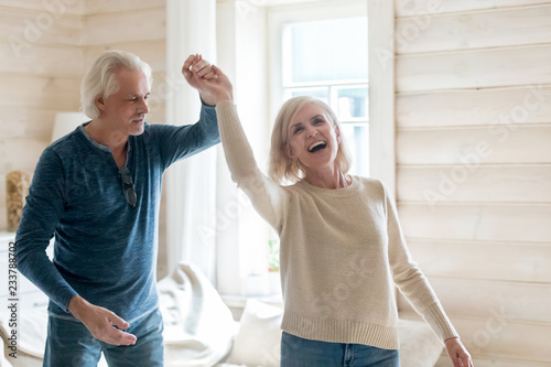 Happy senior husband and wife have fun spending time in country house together, smiling aged couple dance swirling and swaying at home, excited elderly man and woman waltz turning around