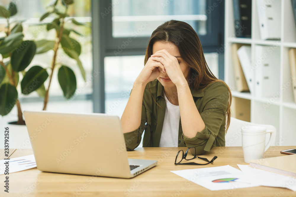 Feeling tired and stressed. Frustrated young woman keeping eyes closed and massaging nose while sitting at her working place in office Feeling tired and stressed. Frustrated young woman keeping eyes closed and massaging nose while sitting at her working place in office