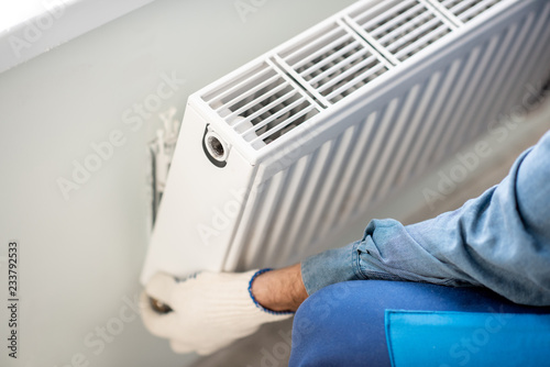 Workman mounting water heating radiator on the white wall indoors, close-up view