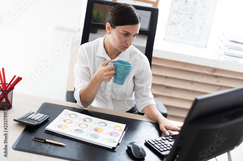 Tableau sur toile A young girl sitting at a table in the office, holding a Cup and working with a computer and documents