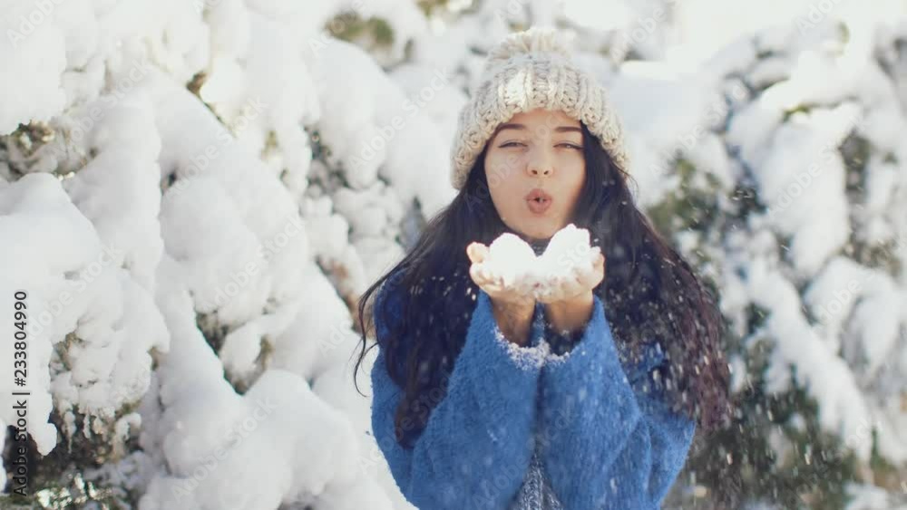 Happy winter fun woman blowing snow in slow motion. Outdoor lifestyle girl playing in snow outside laughing in blue coat, hat, scarf. Happy young woman with winter clothes.