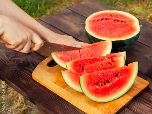 Women's hands cut with a knife into slices of ripe watermelon on a wooden table