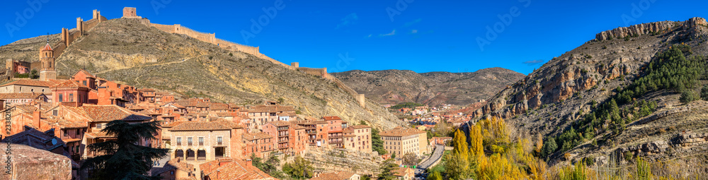 A panoramic photo taken from inside the town of Albarracin Spain where the ancient wall can be seen along with the rooftops, and the mountains and golden trees of autumn at visible under a blue sky