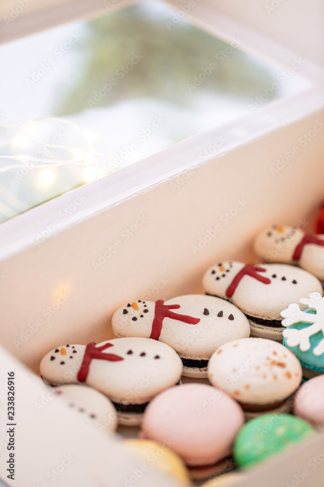 Fototapeta premium Variety of sweet macaroons in white box. The branches of spruce on bokeh background. Modern european French cuisine. Christmas theme, Merry Christmas card. New year mood. lights of garland bulbs