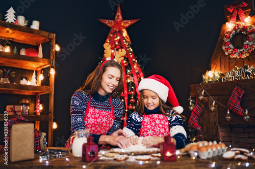 Merry Christmas and Happy Holidays. Family preparation holiday food. Mother in sweater, Christmas deer antlers and daughter in Santa hat cooking cookies and having fun.