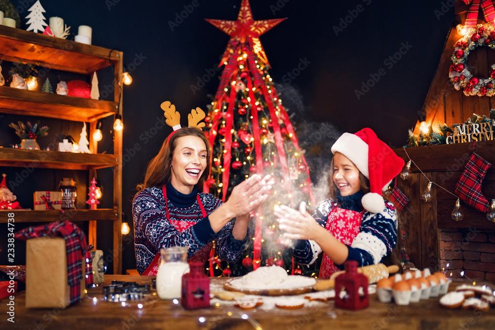 Merry Christmas and Happy Holidays. Family preparation holiday food. Mother in sweater, Christmas deer antlers and daughter in Santa hat cooking cookies and having fun. Merry Christmas and Happy Holidays. Family preparation holiday food. Mother in sweater, Christmas deer antlers and daughter in Santa hat cooking cookies and having fun.