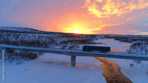 AERIAL: Semi truck crossing the bridge above icy river in the winter at sunset