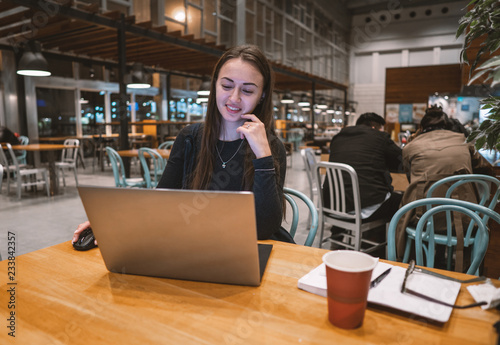 Young, beautiful girl working with laptop and drinking coffee at a wooden table in an empty airport terminal cafe. she is smiling
