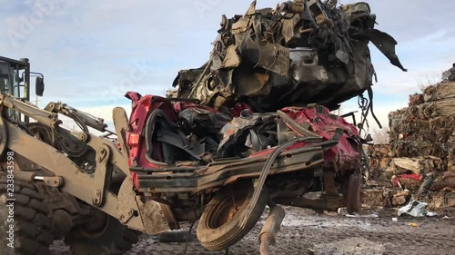 Crushed cars in junk yard are lifted up by forklift and pass by at sunset towards salvage piles in the distance. 