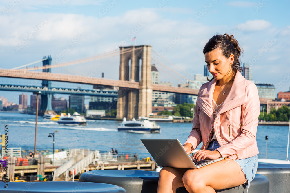 Young American Woman traveling, working in New York, wearing light pink leather jacket, blue Denim shorts, sitting by river, working on laptop computer. Brooklyn, Manhattan bridges, boat on background