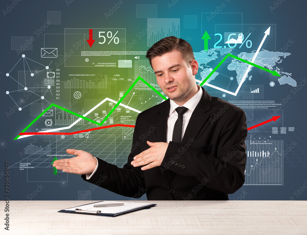 Young handsome businessman sitting at a desk with stocks and progress charts behind him Young handsome businessman sitting at a desk with stocks and progress charts behind him