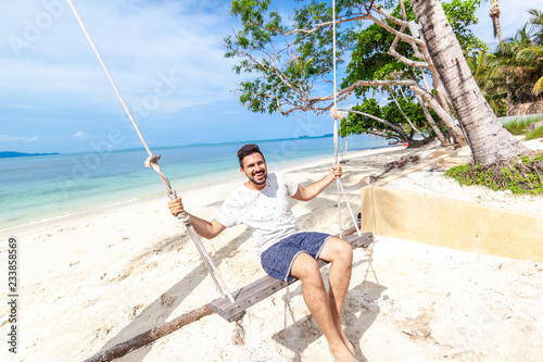 Young attractive happy mix raced man swinging on a swing on the shore of a bright tropical sea, vacations and travel