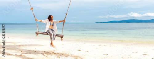 Beautiful young woman in a white shirt swinging on a swing on the beach, against the backdrop of a paradise landscape with turquoise water