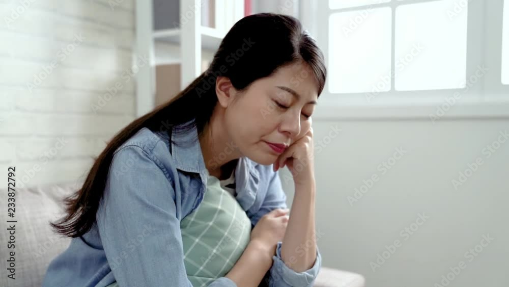 micromotion of an upset asian female shaking head sitting on the couch. young office lady lost her job staying at home and feel depressed. attractive thoughtful woman daydreaming in the cozy room. micromotion of an upset asian female shaking head sitting on the couch. young office lady lost her job staying at home and feel depressed. attractive thoughtful woman daydreaming in the cozy room.