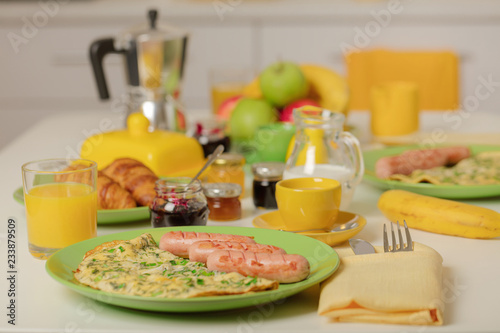Breakfast time. Omelet with greens and sausages. Croissants and orange juice, jam. Coffee with cream or milk. Fruits - bananas, red and green apples.