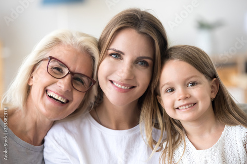 Headshot portrait of three 3 generations family, smiling grandmother, grown young daughter and child girl looking at camera, happy kid granddaughter, mother and old aged grandma posing together