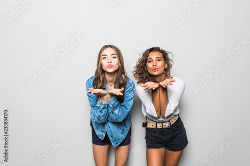 Two multiethnic girls blow airkiss at camera, wears trendy shades, summer clothing, express love or say goodbye on distance, pose against gray background. People, style and diversity concept photo