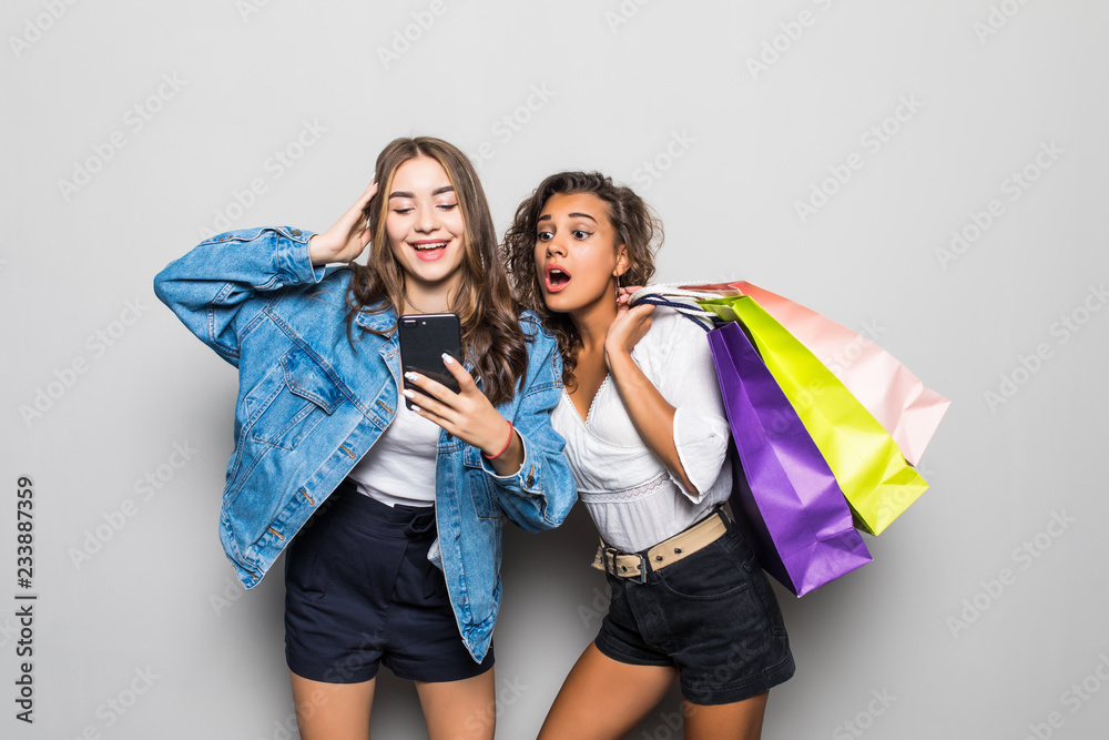 Portrait of surprised young two ladies friends with bright makeup lips standing over grey wall and posing with shopping bags using mobile phone. Portrait of surprised young two ladies friends with bright makeup lips standing over grey wall and posing with shopping bags using mobile phone.