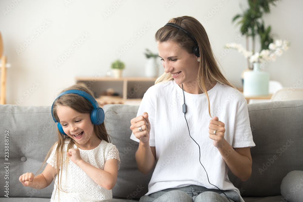 Happy young mother and funny child daughter having fun laughing dancing sitting on couch listening to music in earphones together, smiling mom and kid wearing headphones enjoy favorite songs at home Stock Happy young mother and funny child daughter having fun laughing dancing sitting on couch listening to music in earphones together, smiling mom and kid wearing headphones enjoy favorite songs at home Stock