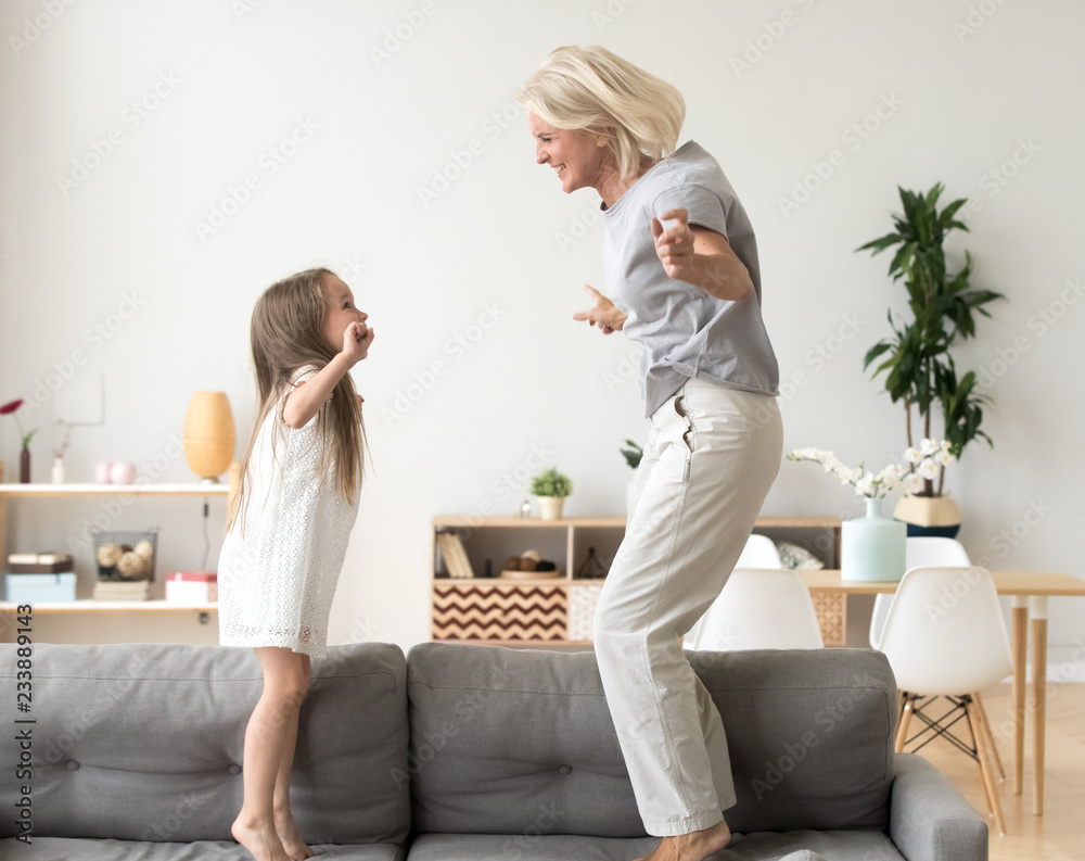 Cute little girl having fun playing with smiling grandmother jumping on couch together, happy granny and active kid grandchild dancing on sofa, grandma and granddaughter laughing playing at home - obrazy, fototapety, plakaty 