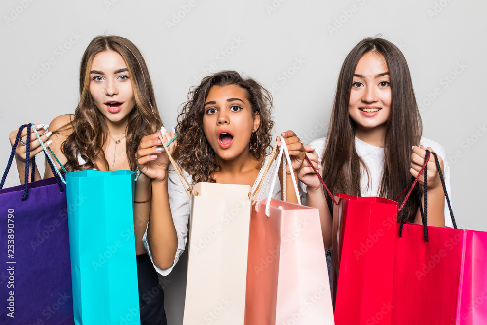 Three beautiful shocked women looking inside shopping bags isolated over gray background. Black friday sales concept. Shopping time.