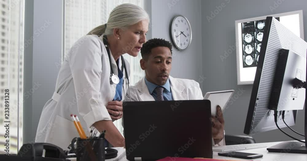 Millennial male doctor using tablet and looking at computer screen with colleague. Two medical doctors working inside office using technology