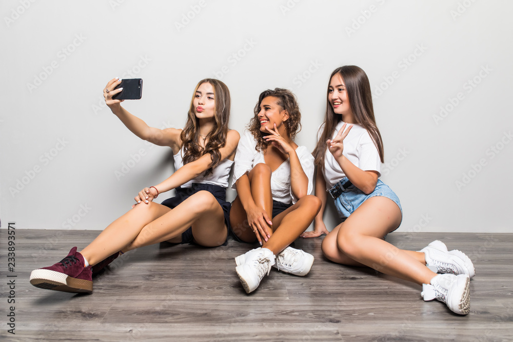 Portrait of multiethnic three young cute pretty girls friends make selfie by mobile phone sitting isolated over white background .