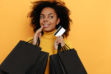 Black friday. Shopping. Afro American girl in yellow sweater is holding shopping bags and a credit card and smiling, on a yellow background