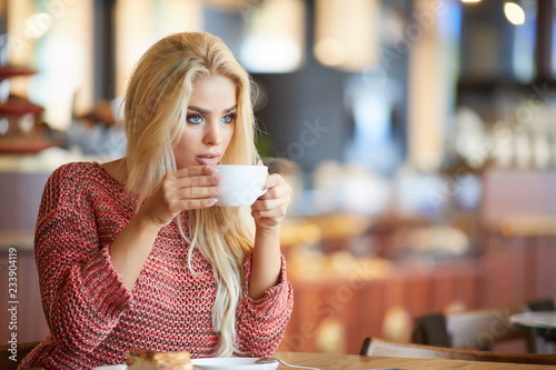 Blonde woman looking through window while drinking tea sitting in cafe