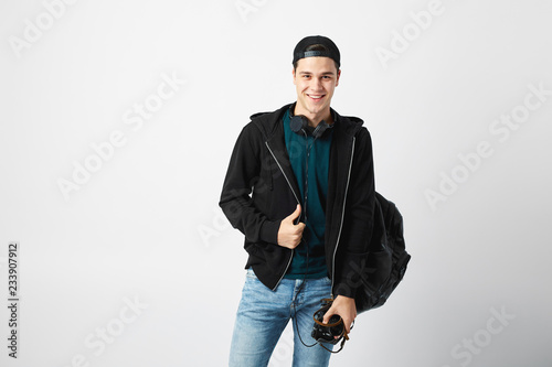 Smiling guy with a black backpack on his shoulder dressed in a dark green t-shirt, jeans, sweatshirt and a cap holds a camera in his hand on a white background