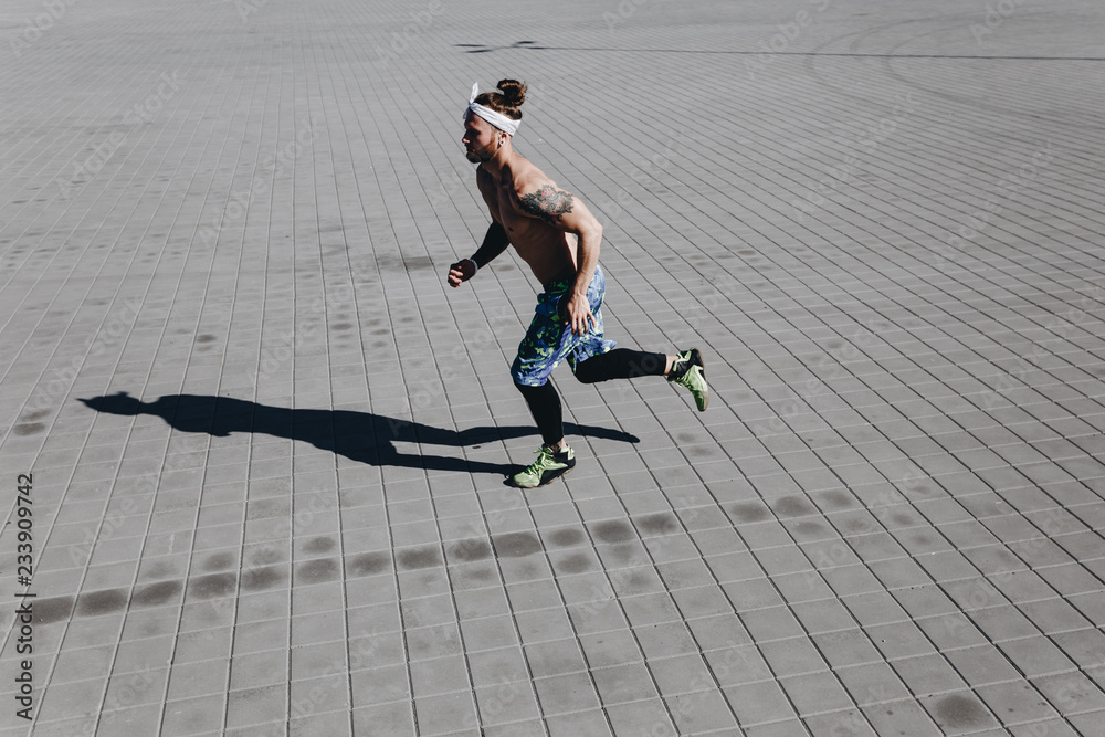 Obraz premium Young athletic man with a naked torso with tattoos and headband on his head dressed in the black leggings and blue shorts runs on paving slabs on a warm sunny day
