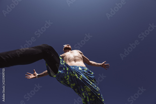 Fototapeta Naklejka Na Ścianę i Meble -  Young athletic man with a naked torso with headband dressed in the black leggings and blue shorts is jumping high on the background of blue sky on a warm sunny day