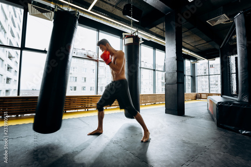 Guy in red boxing gloves with a naked torso dressed in the black shorts hits punching bag in the gym against the background of panoramic windows