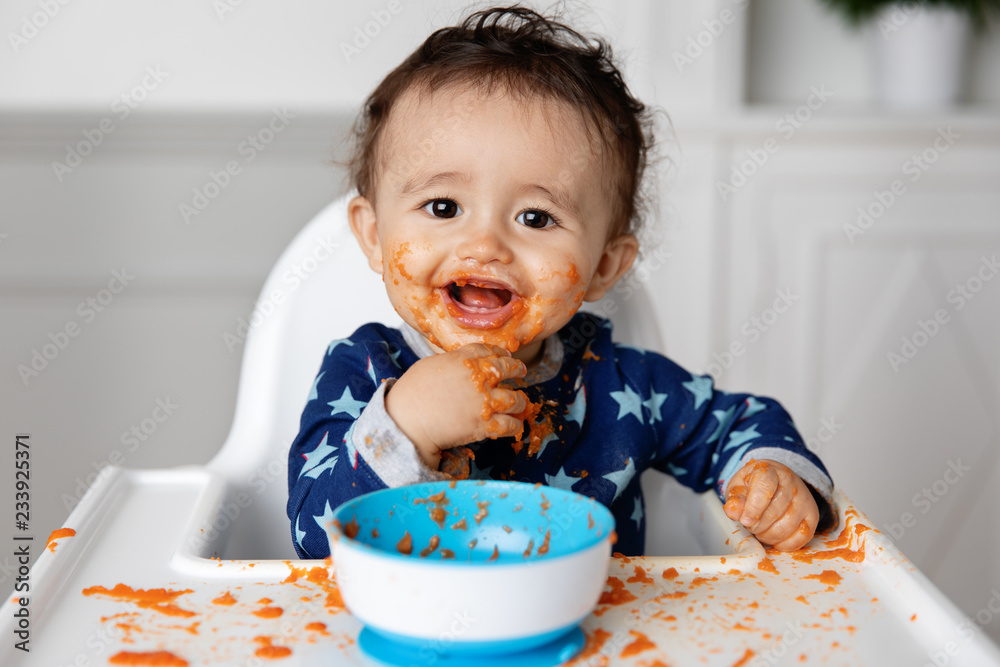 Smiling baby making a mess with carrot puree in high chair Smiling baby making a mess with carrot puree in high chair