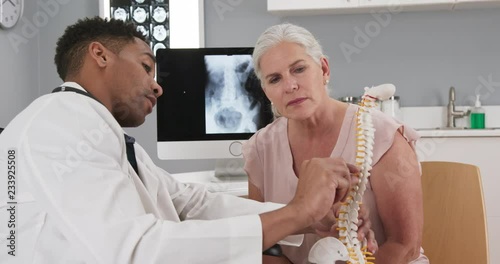 Senior female patient consulting with young doctor about back injury. Close up of black doctor holding model of spinal cord and explaining to elderly woman source of her pain