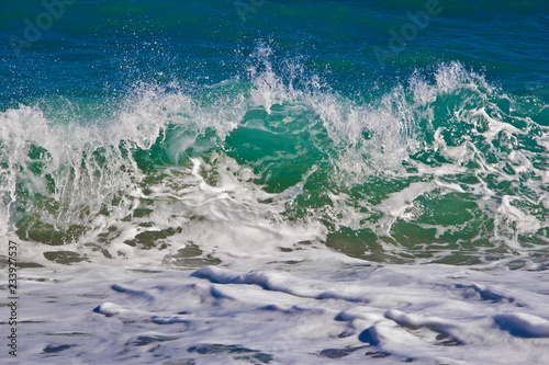 A large beautiful emerald wave crashes near the shore. Small splashes of fun sparkle in the sun on a bright sunny day. Sea foam spread on the shore