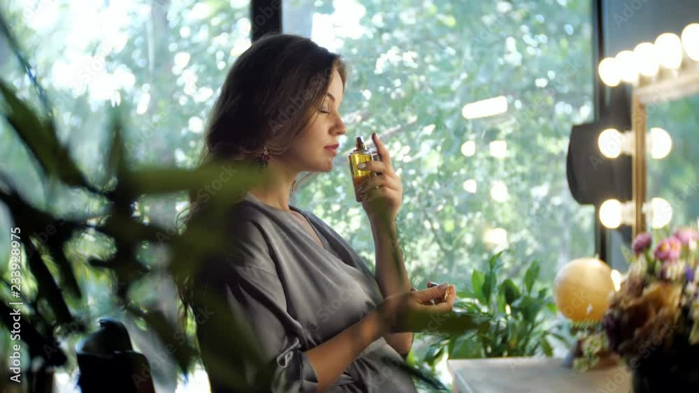 Side view of elegant brunette in silk robe sitting against mirror and spraying herself with perfume