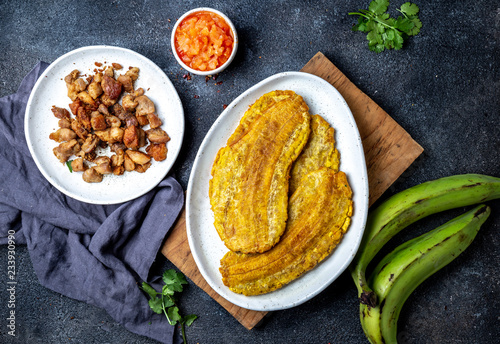 COLOMBIAN CARIBBEAN CENTRAL AMERICAN FOOD. Patacon or toston, fried and flattened whole green plantain banana on white plate with tomato sauce and chicharron Black background, top view photo