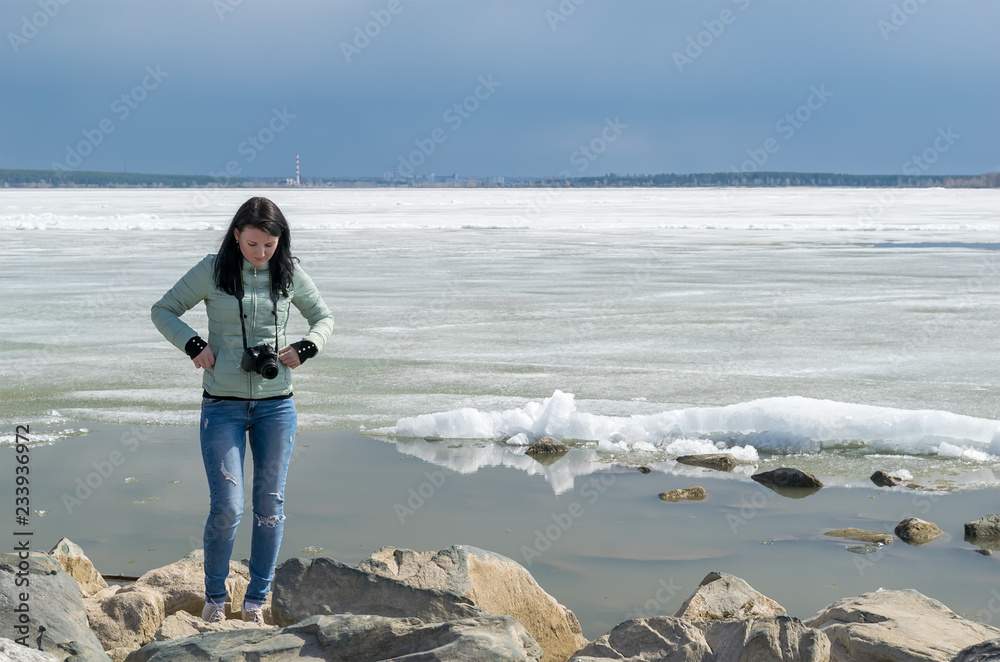 spring, the girl in warm jacket with a camera stands on the shore of the lake and tries to go over the stones