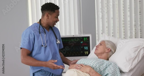 Young black nurse talking to senior woman resting in hospital bed. Male RN assisting elderly female patient lying down in bed