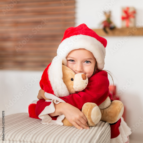 Cute young girl wearing santa hat hugging her christmas present, soft toy teddy bear. Happy kid with xmas present, smiling and looking at camera.
