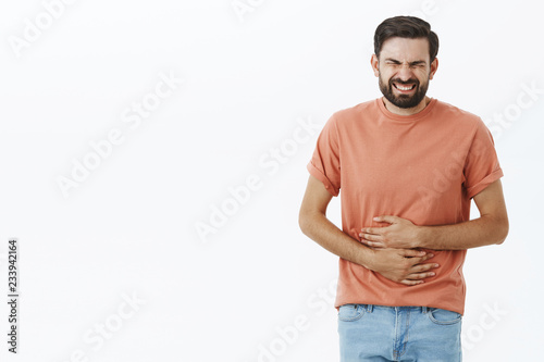 Guy regrets eating fast food feeling pain in stomach clenching teeth and closing eyes from pain touching belly suffering disorder or stomachache waiting for ambulance posing against gray background photo